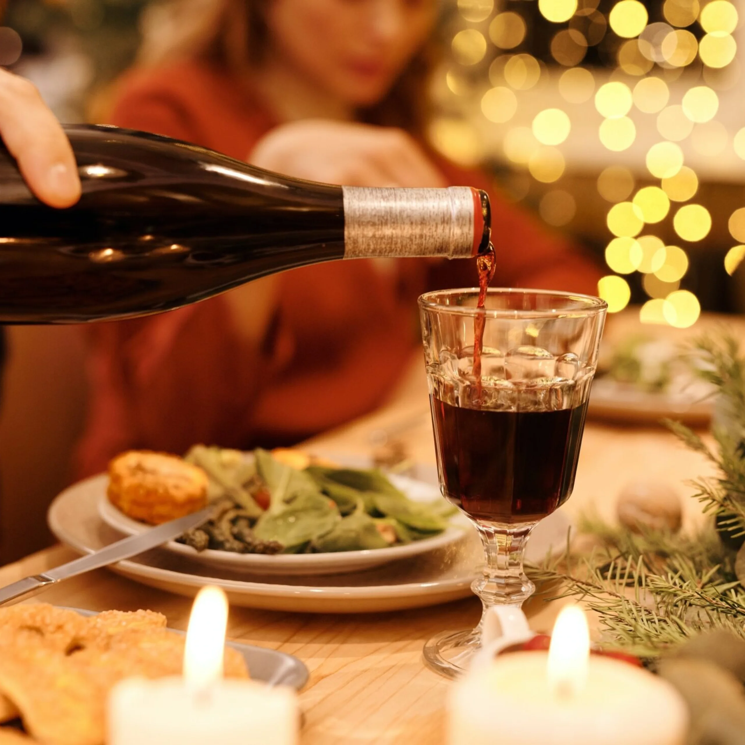 someone pouring red wine from a bottle into a glass with christmas lights in the background