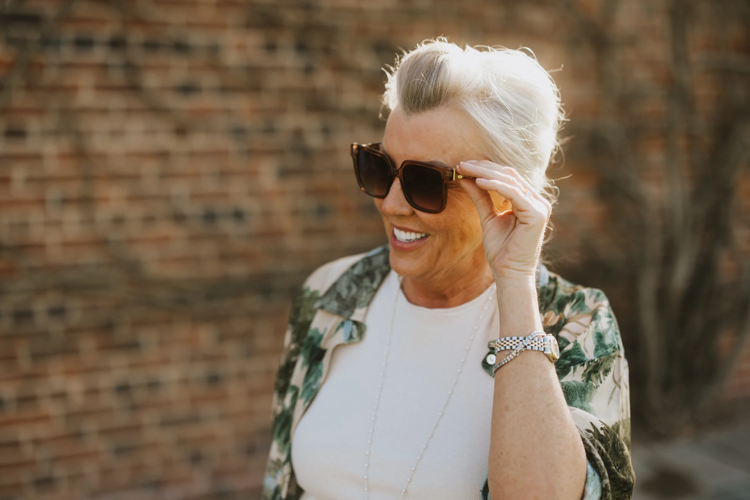 A woman poses in the sun holding the arm of her sunglasses