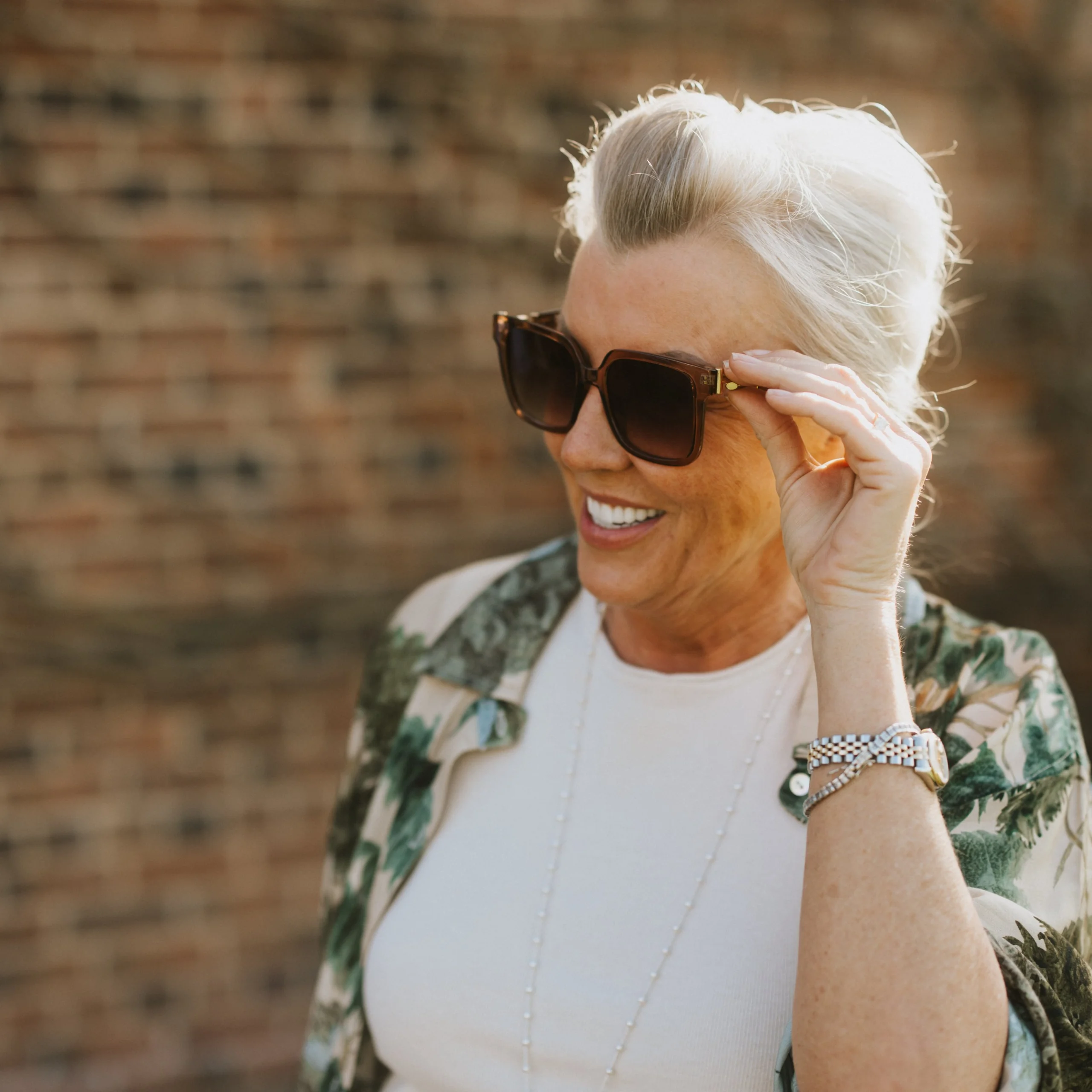 A woman poses in the sun holding the arm of her sunglasses