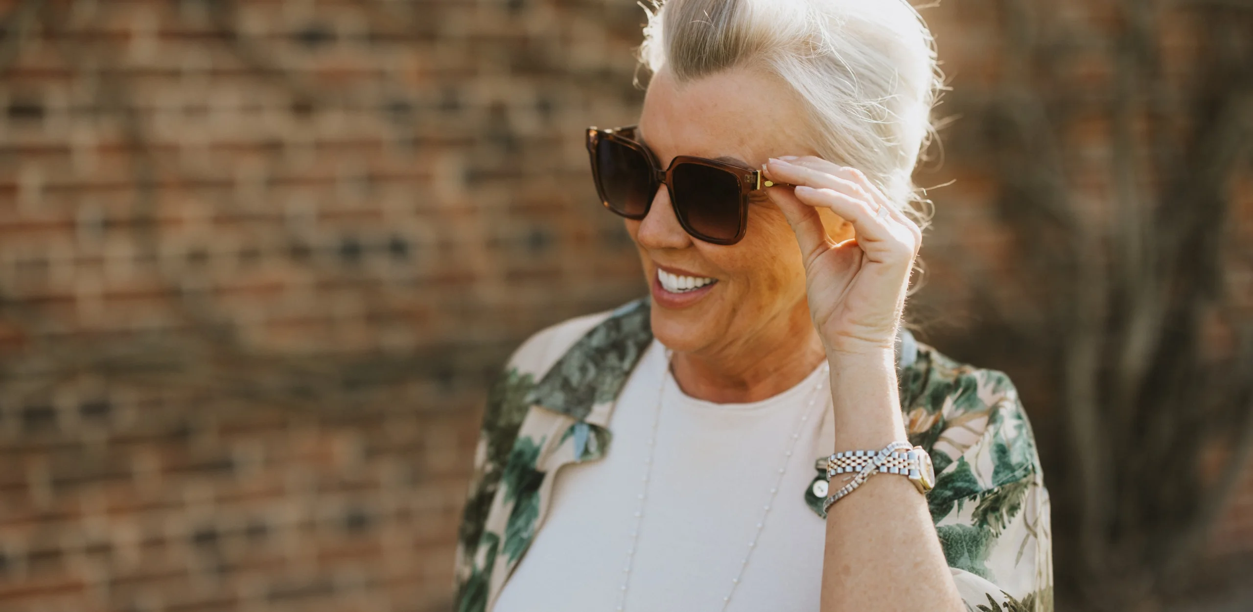 A woman poses in the sun holding the arm of her sunglasses