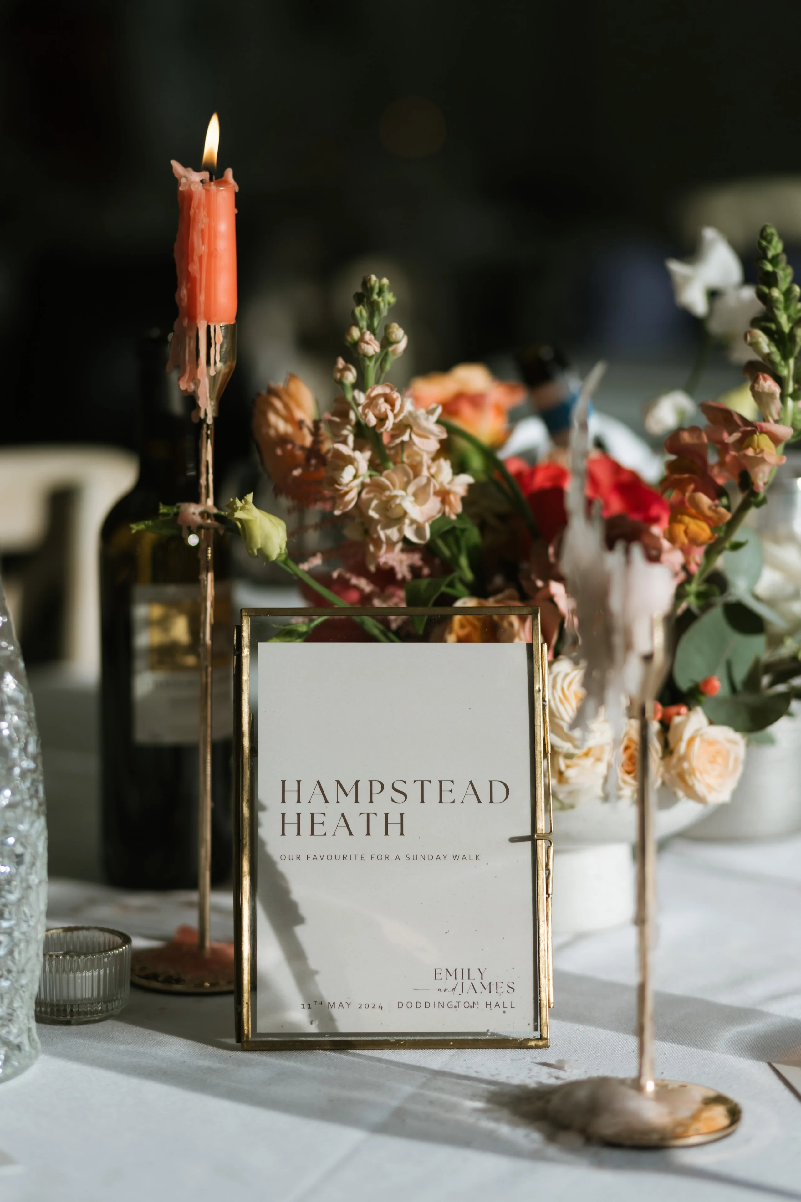 a wedding table setup with a table name and seasonal orange and peach flowers with an orange dinner candle in a gold holder
