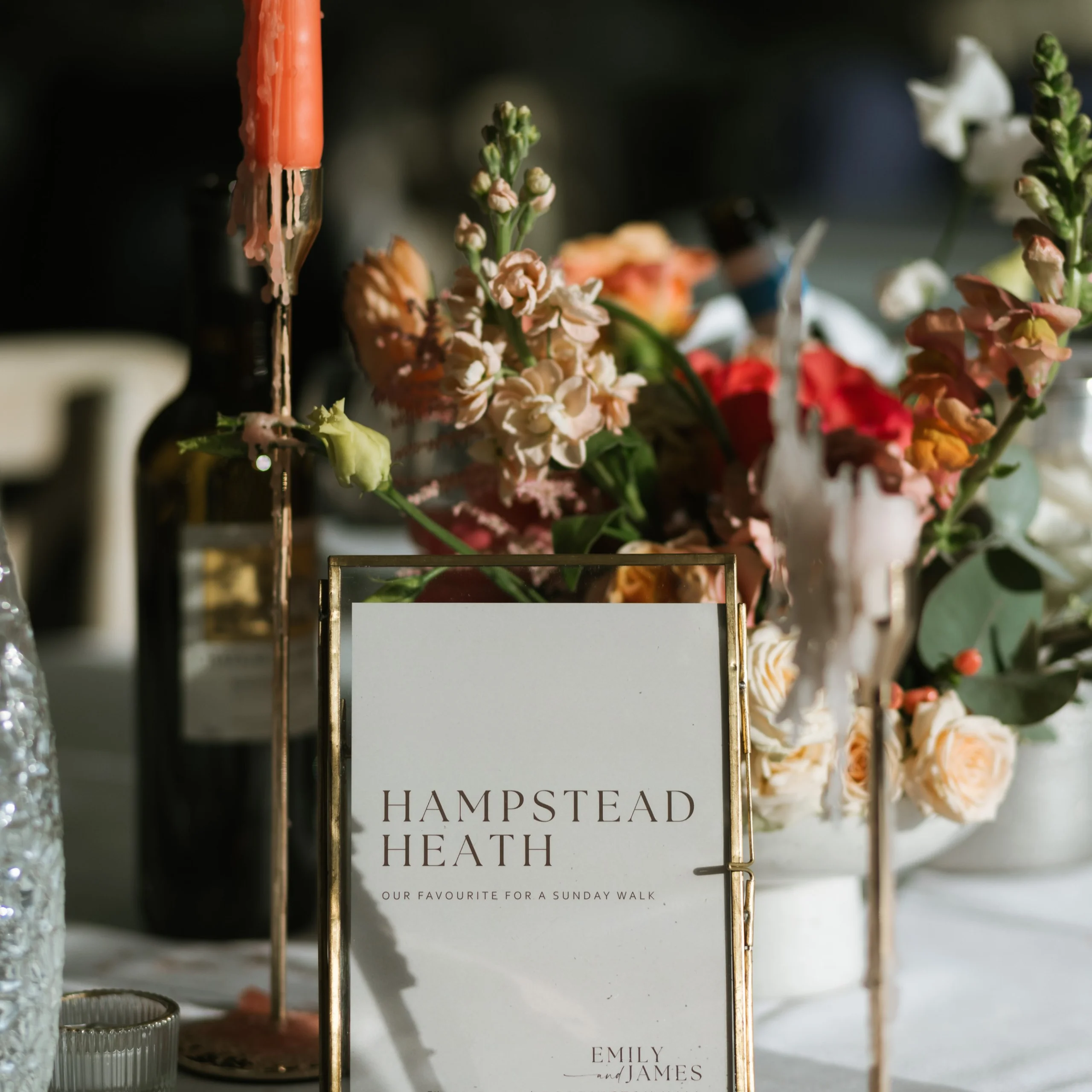 a wedding table setup with a table name and seasonal orange and peach flowers with an orange dinner candle in a gold holder