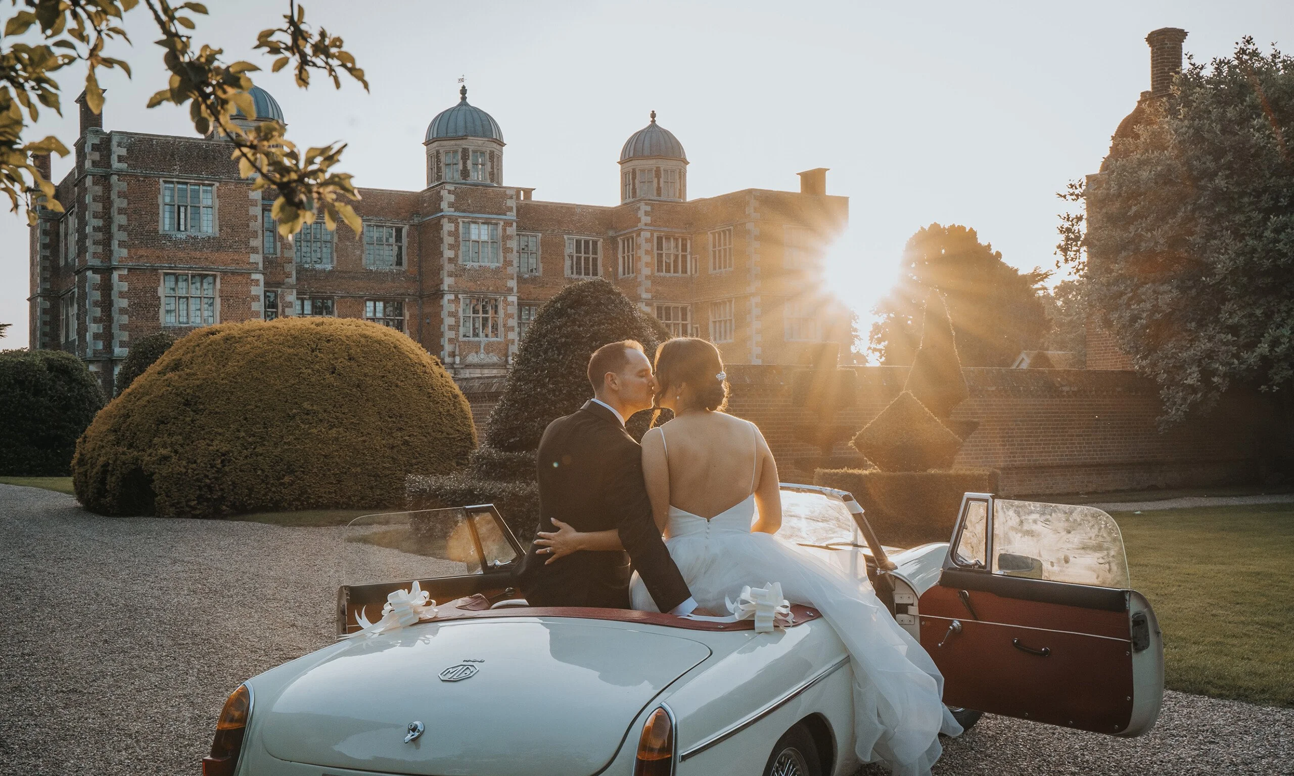 a bride and groom sat on the back of a vintage car kissing with doddington hall in the background