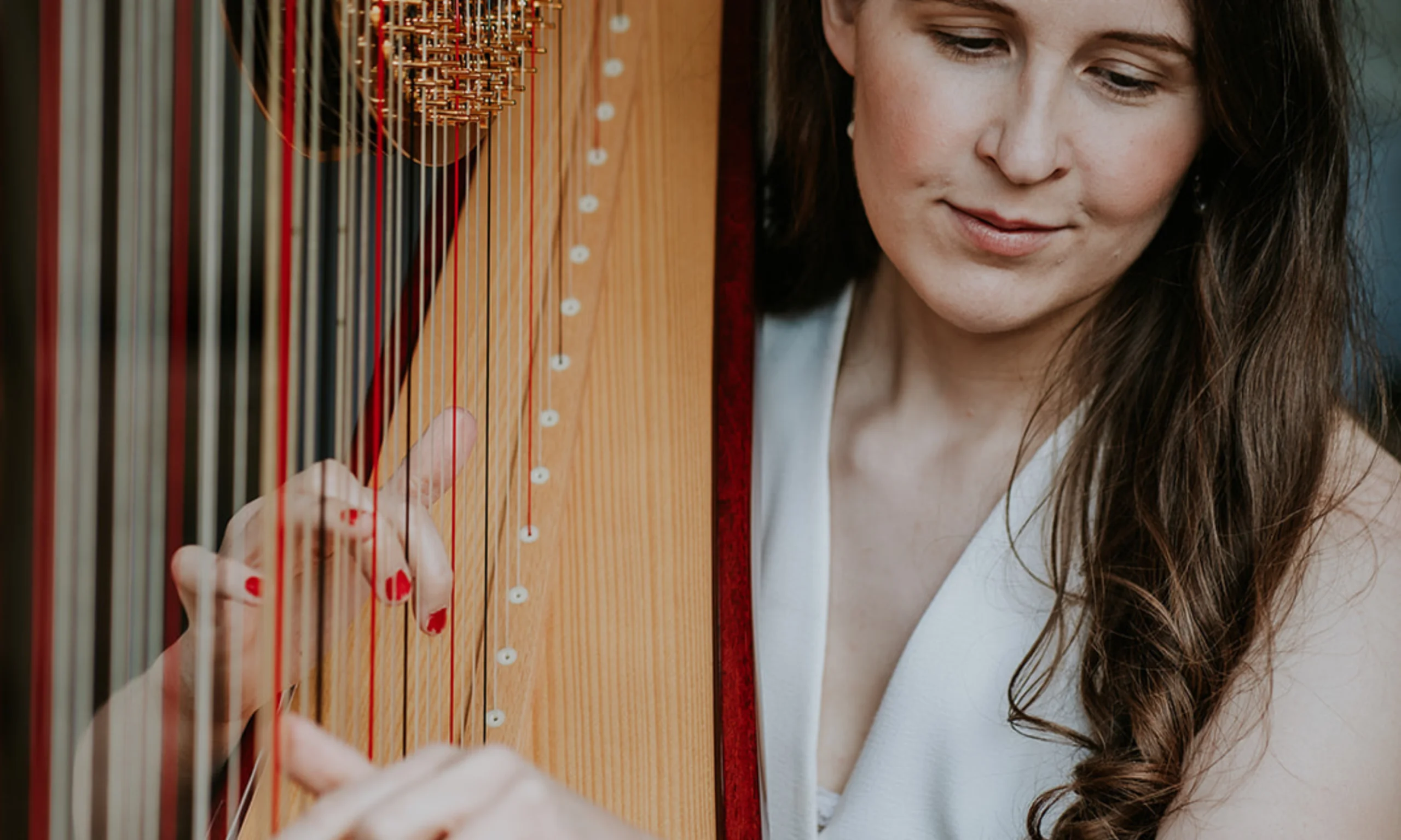 a lady wearing a white waistcoat strumming a harp with red nail varnish on her finger nails